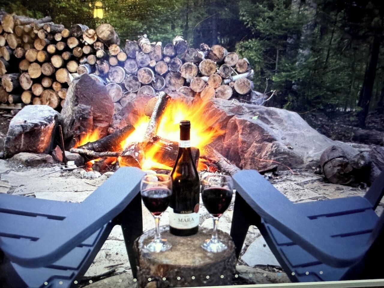 A campfire with logs stacked in the background, a bottle of wine, and two glasses on a table in the foreground.