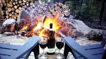 A campfire with logs stacked in the background, a bottle of wine, and two glasses on a table in the foreground.