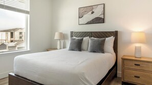 A neatly made bed with a wooden headboard, two bedside lamps, and a view of buildings through a window. - Landing | Amazing 2BD, Clubhouse, Gym (Longmont)