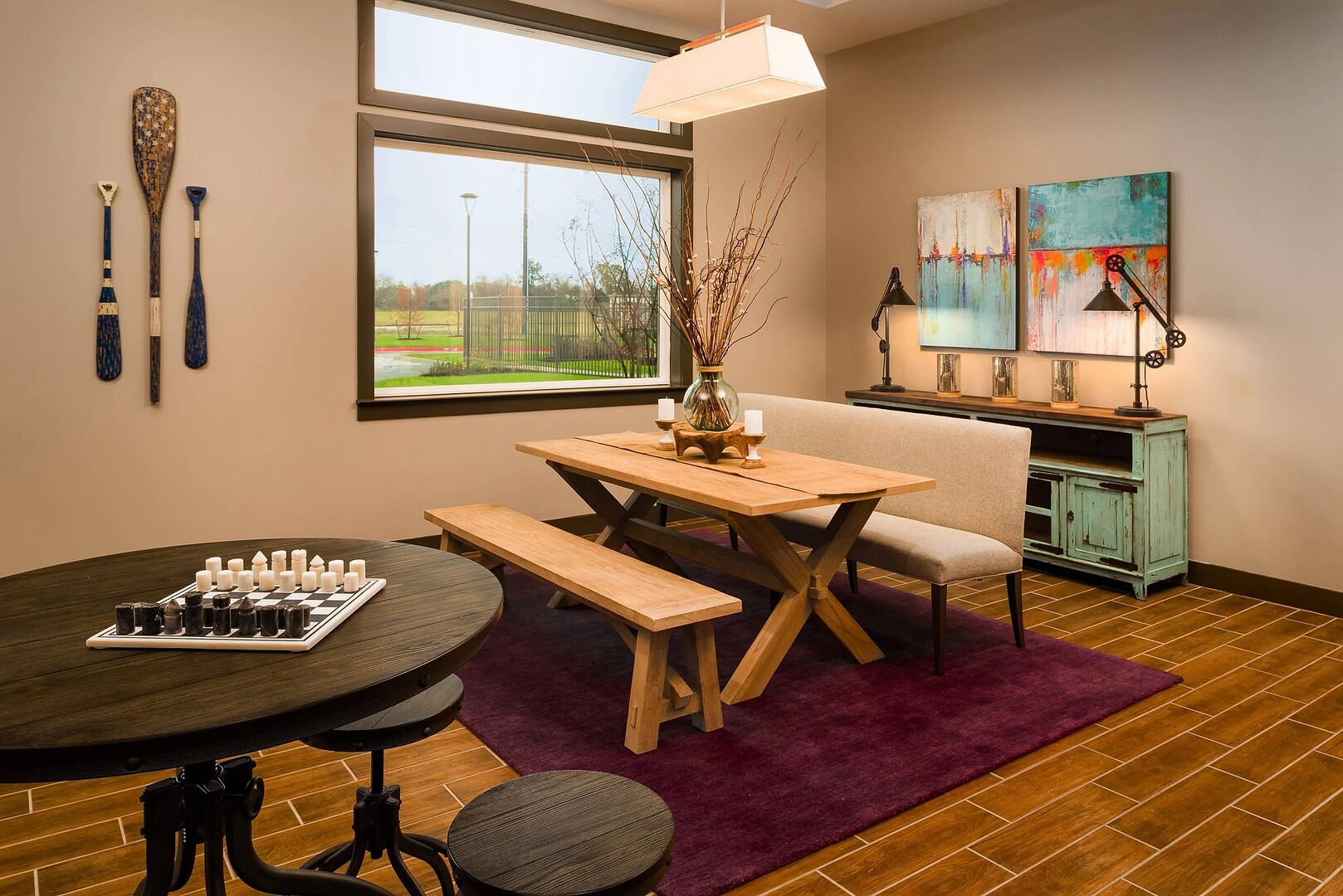 A cozy room with a wooden table, bench, and a chessboard. There's a window with a view of a park, a vase with branches, and a painting on the wall.