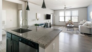 A modern kitchen with granite countertops, a dishwasher, and a dining area with a coffee table and sofa. - Landing | Chic 1BD in Eastpoint (Baytown)