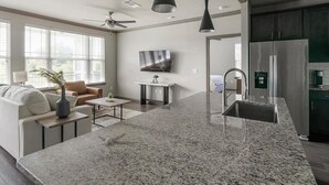 A modern kitchen with a granite countertop, stainless steel appliances, and a dining area with a sofa and coffee table. - Landing | Chic 2BD in Eastpoint (Baytown)