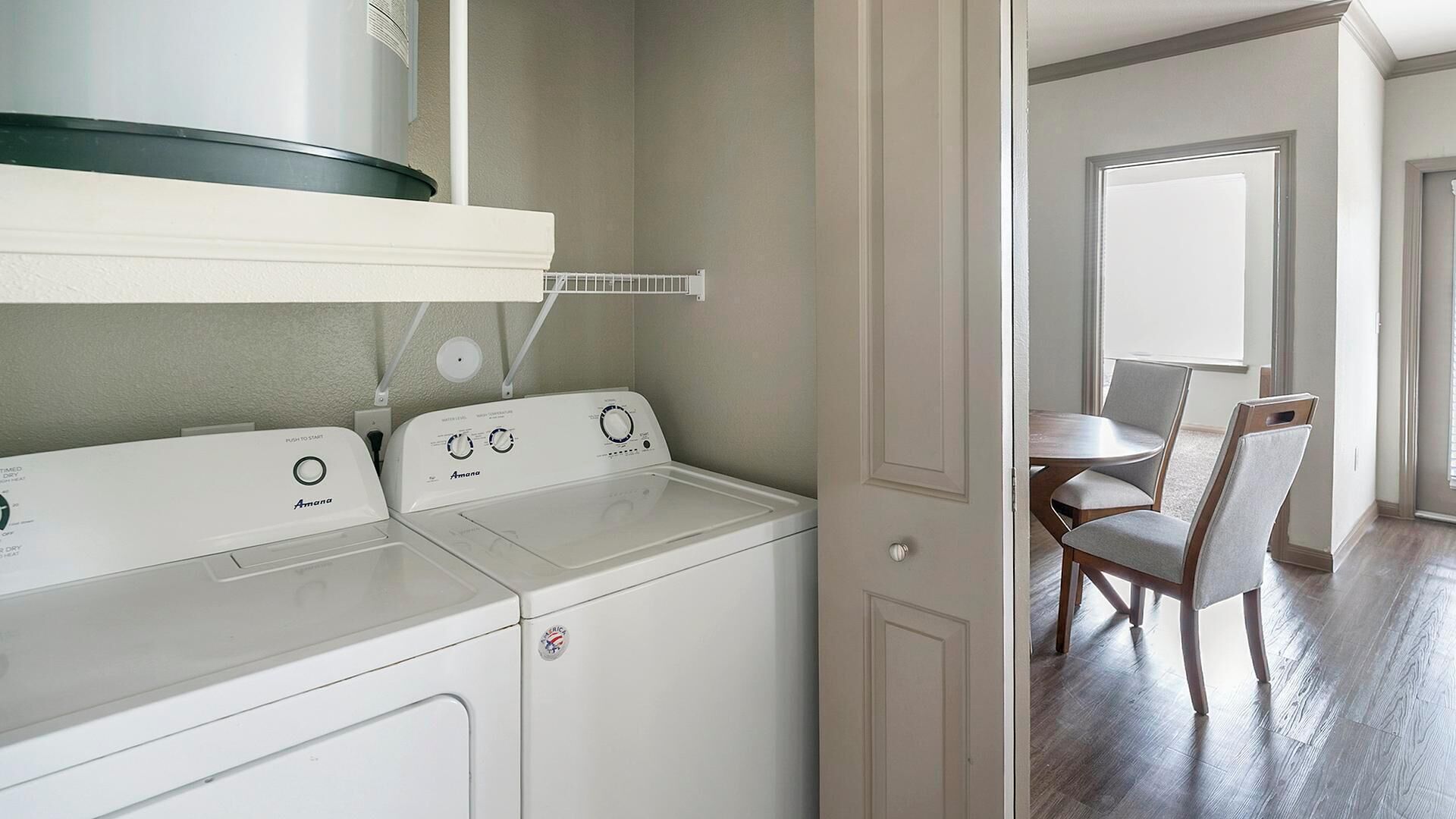 A laundry room with two white Amana washers and dryers, a white shelf above, and a dining area with a table and chairs visible through an open door.