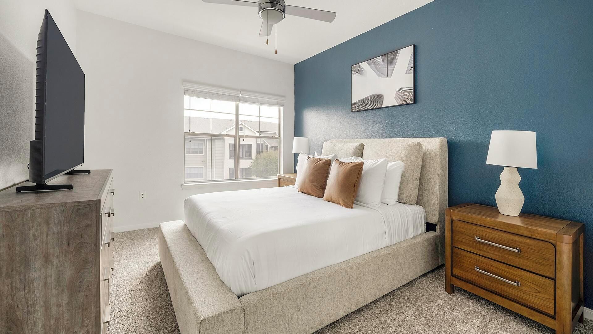 Une chambre à coucher comprenant un lit, une table de chevet, une lampe, un ventilateur de plafond, une fenêtre et un téléviseur.
