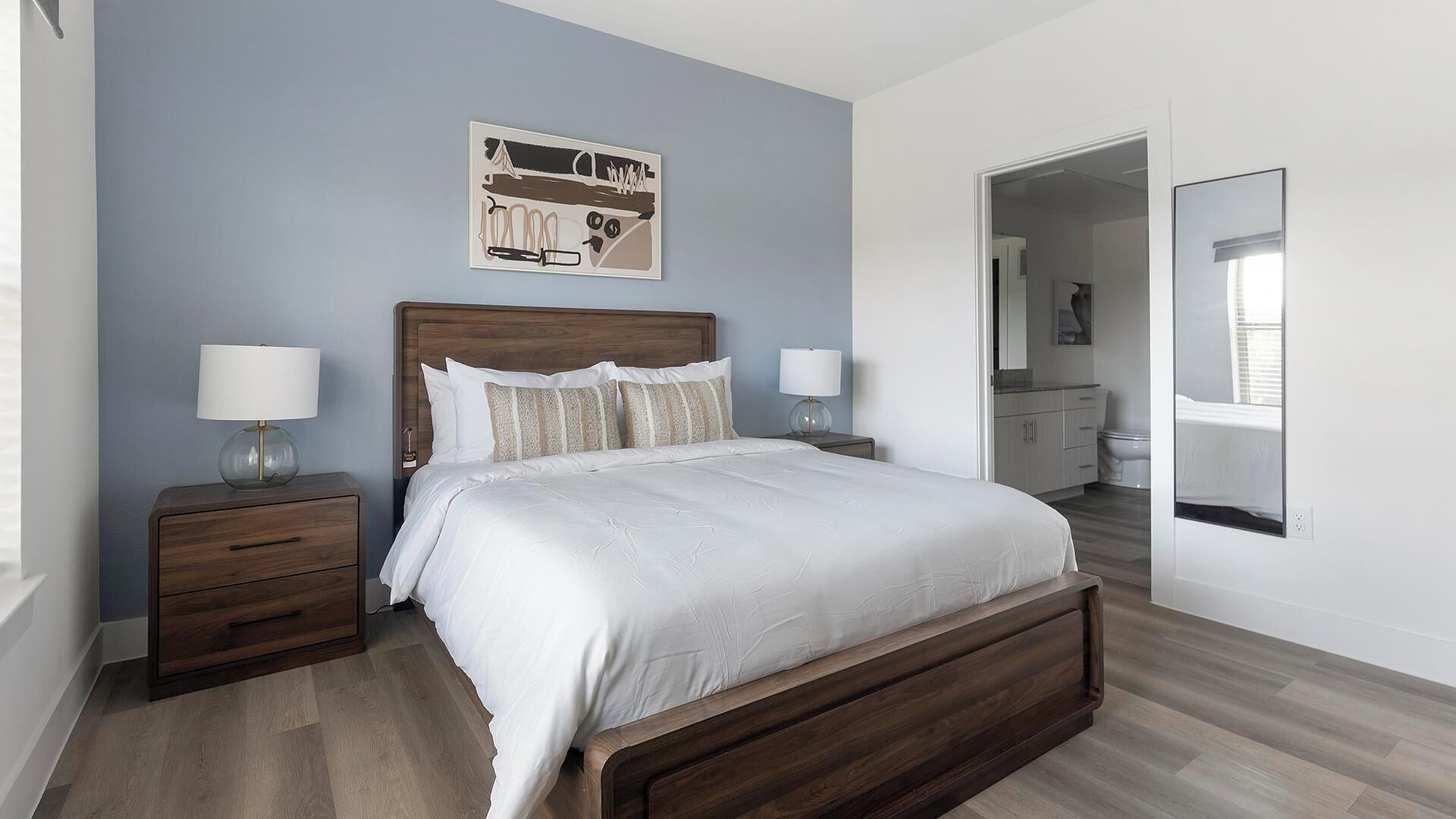 Un dormitorio con una cama de madera, dos mesitas de noche con lámparas, una obra de arte colgada en la pared y un baño visible a través de una puerta abierta.