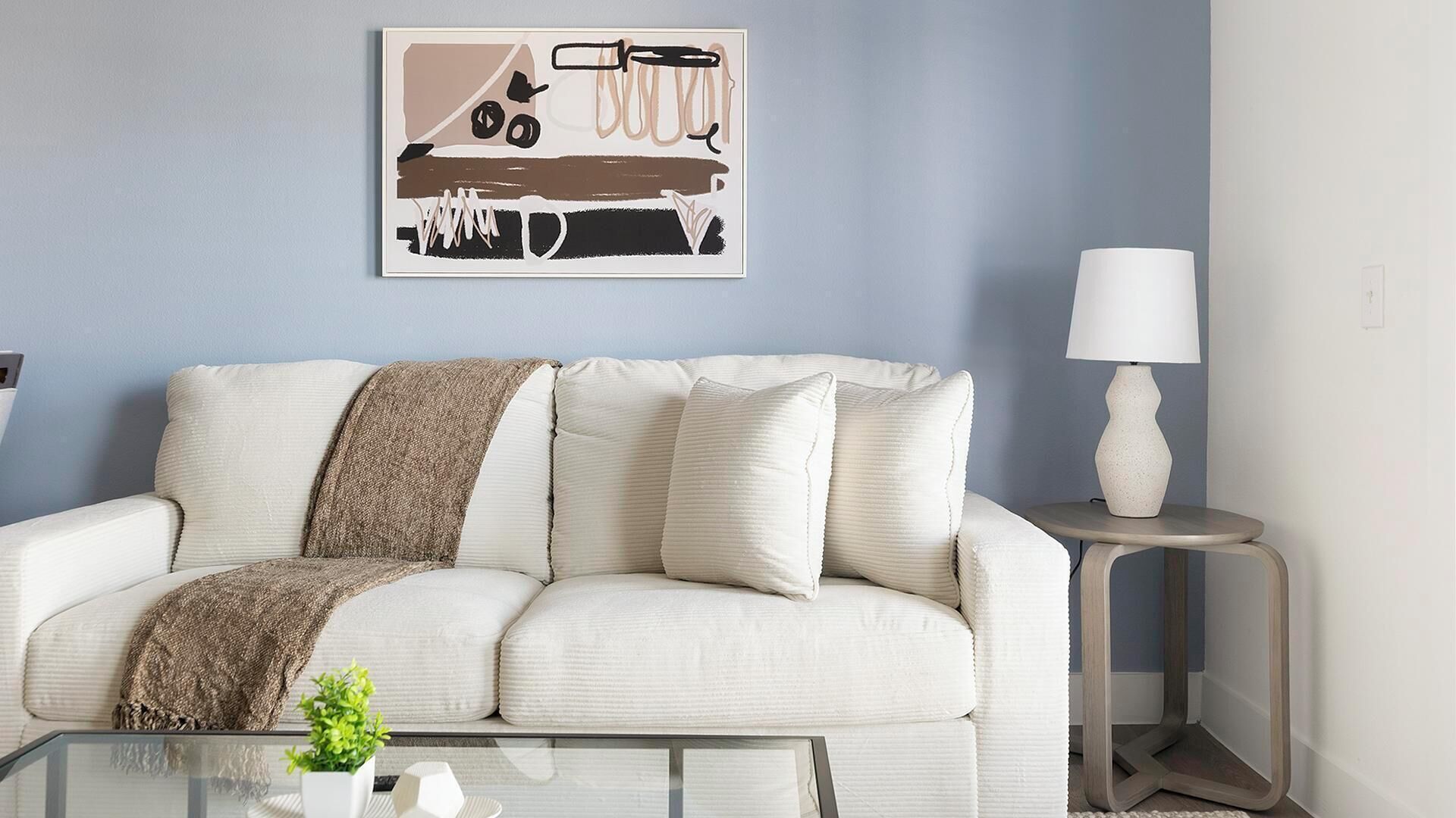 Een woonkamer met een witte bank, een bruin plaid, een glazen salontafel met een plant en een wandlamp.