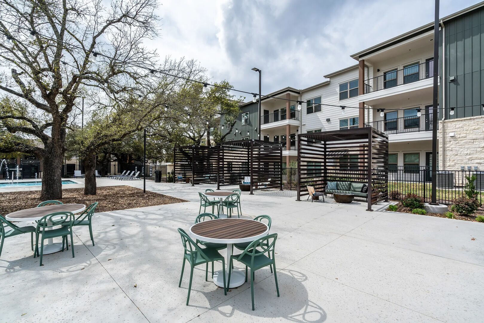 An outdoor patio area with round tables and chairs, a pergola, and a pool in the background.