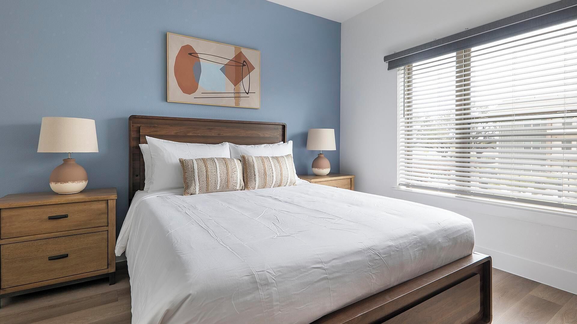 Une chambre à coucher comprenant un lit, deux tables de chevet, une commode, une fenêtre avec stores et une œuvre d’art accrochée au mur.