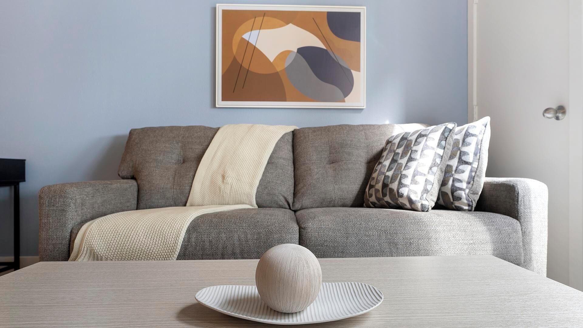Ein Wohnzimmer mit grauem Sofa, weißer Decke, gemusterten Kissen, einem runden Dekorationsobjekt auf einem Tisch und einem gerahmten abstrakten Gemälde an der Wand.