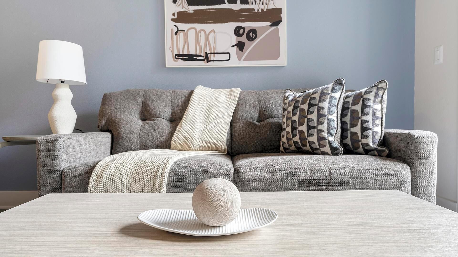 Ein modernes Wohnzimmer mit einem grauen Sofa, gemusterten Kissen, einer weißen Lampe und einem strukturierten Tisch mit einer dekorativen Kugel.