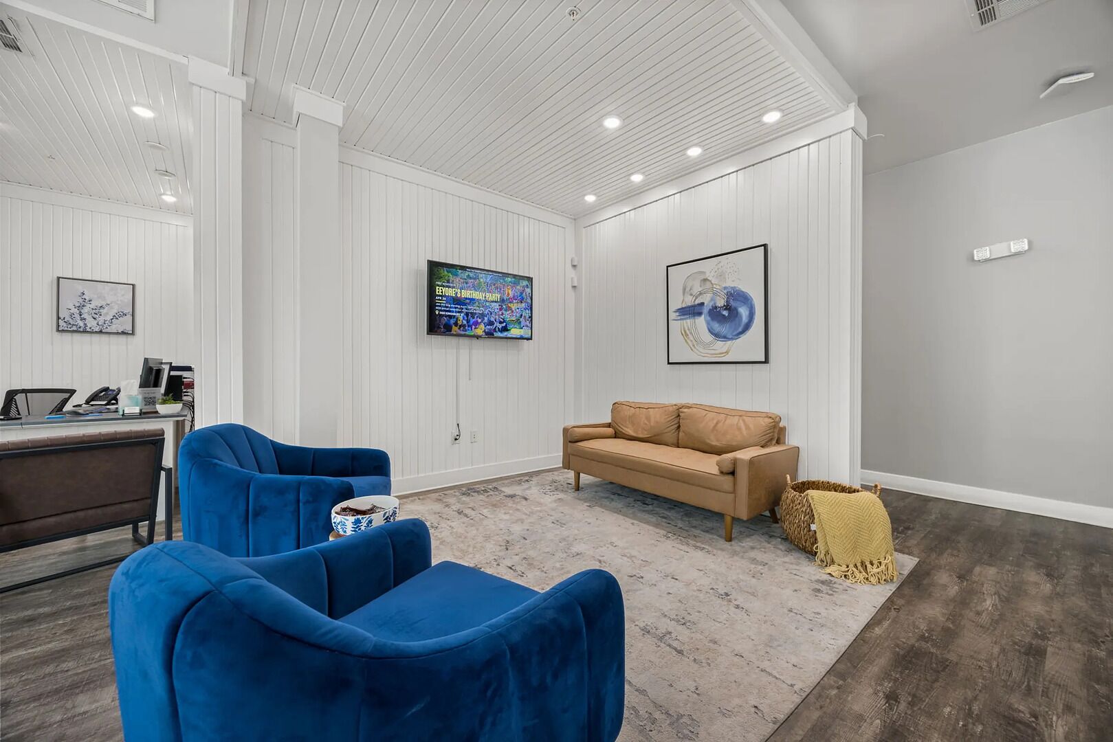 A modern, well-lit room with a desk, two blue chairs, a beige sofa, and artwork on the walls.