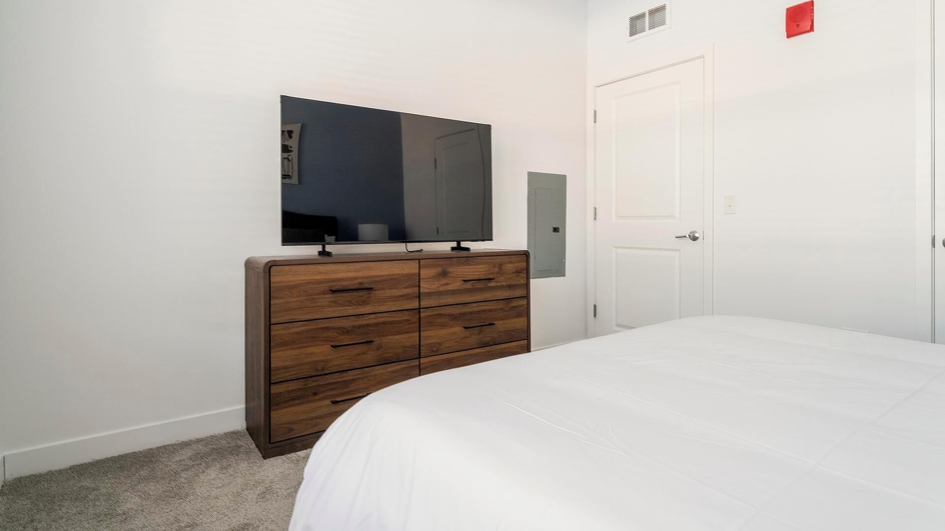 A bedroom with a wooden dresser, a flat-screen TV, a white bed, and a white door.