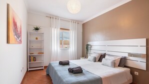 Un dormitorio con cama, cabecera blanca, mesita de noche, estantería y ventana con cortinas.