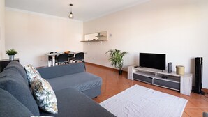 Una sala de estar con un sofá gris, un televisor de pantalla plana, una mesita con sillas, una planta en maceta y un ventilador de pie.