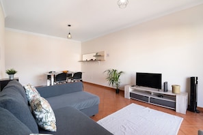 Ein Wohnzimmer mit grauem Sofa, einem Flachbildfernseher, einem kleinen Tisch mit Stühlen, einer Topfpflanze und einem Standventilator.