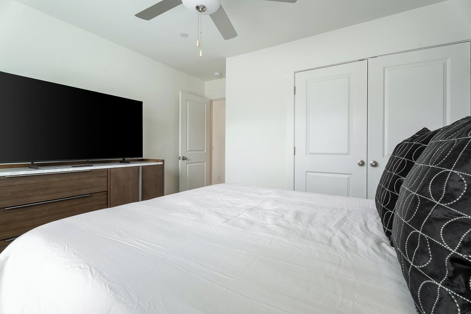 Une chambre à coucher avec un lit, un téléviseur posé sur une commode en bois, un ventilateur de plafond et deux portes blanches.