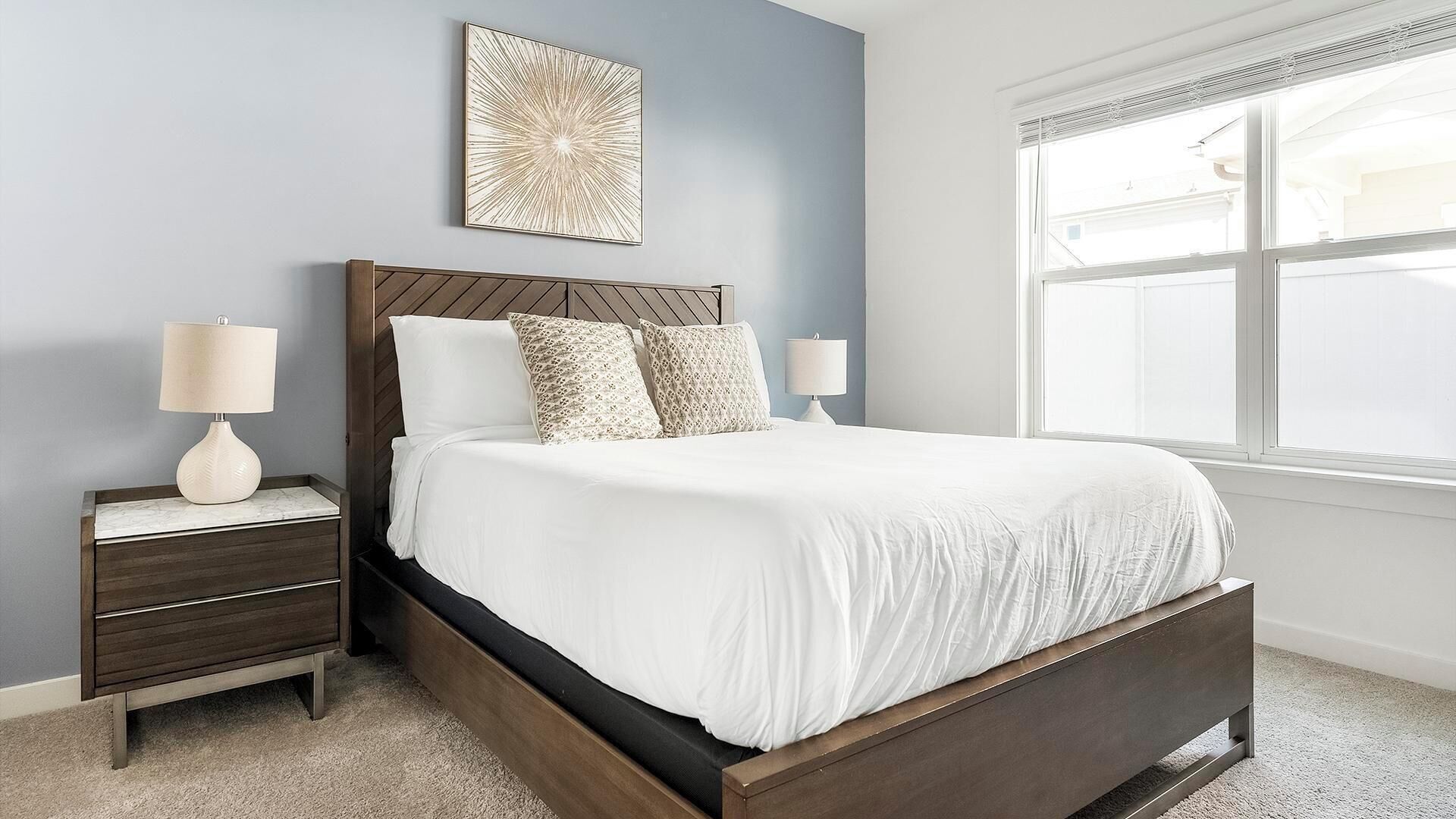 A bedroom with a wooden bed frame, white bedding, two bedside tables with lamps, a wall art piece, and a large window.