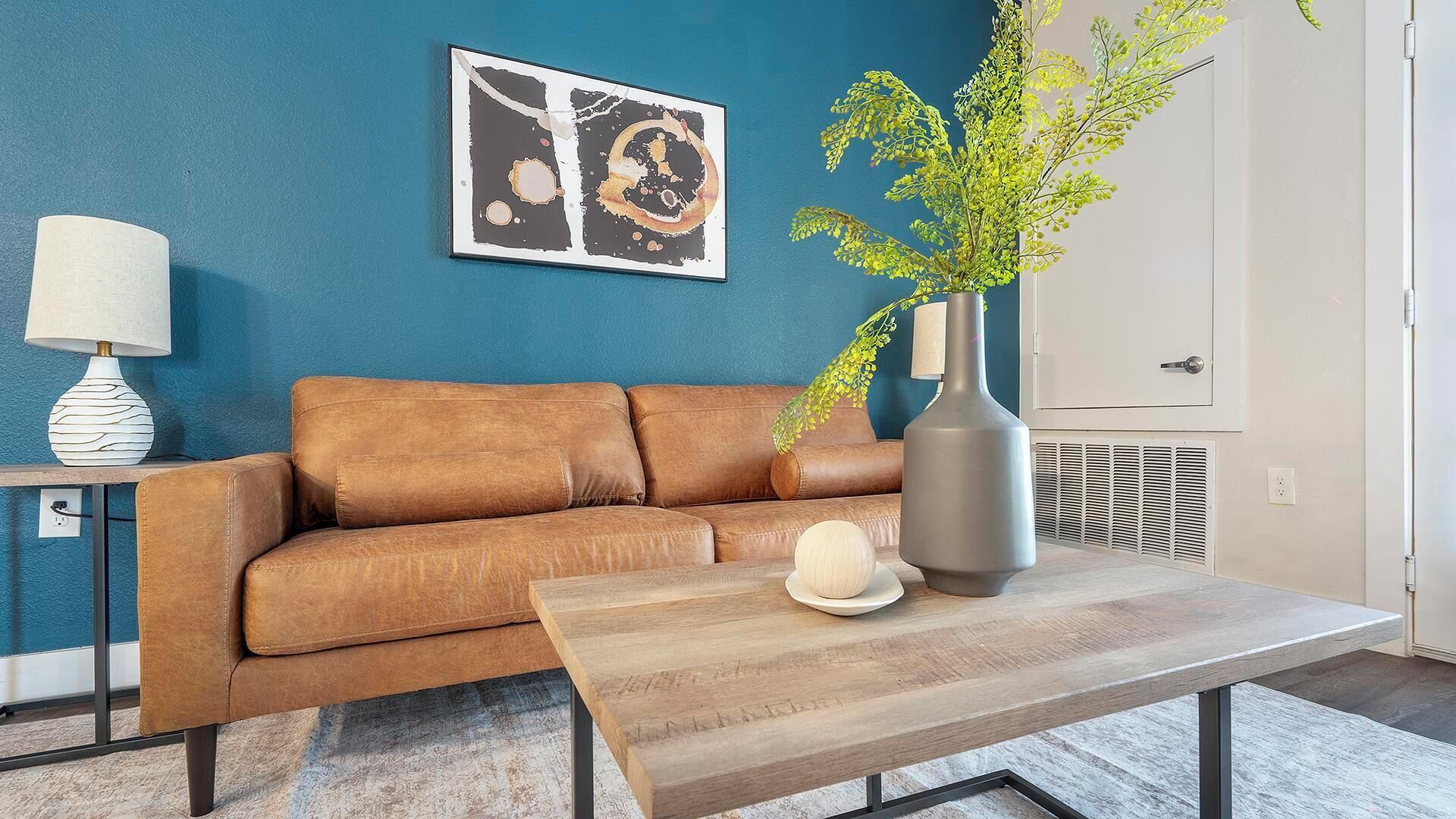 A modern living room with a brown leather sofa, a wooden coffee table, a vase with a plant, and a framed abstract painting on the wall.
