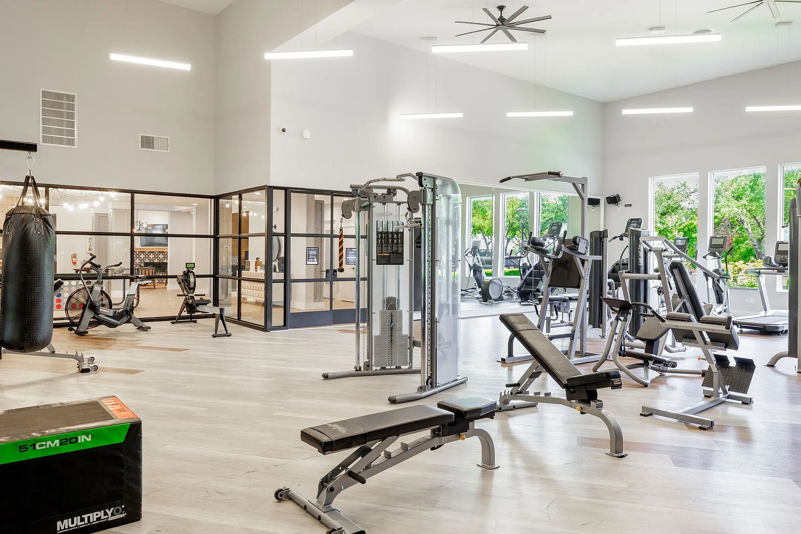 Ett gym med olika typer av träningsutrustning, en boxningssäck och stora fönster med utsikt över grönområden.