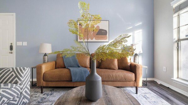 Un salon avec un canapé en cuir marron, un vase gris orné de branches, un plaid à motifs et une œuvre d’art encadrée accrochée au mur.