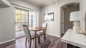 Un coin repas avec une table et des chaises en bois, une grande fenêtre avec des stores, un tableau accroché au mur et un comptoir en marbre blanc surmonté d’une lampe.