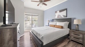 Une chambre à coucher comprenant un lit, des tables de chevet, un téléviseur, un ventilateur de plafond et une fenêtre avec des stores.