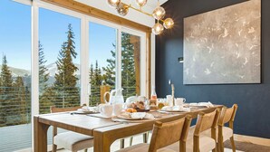 Un comedor con una mesa de madera dispuesta para el desayuno, amplias ventanas con vistas a árboles y montañas, y una lámpara moderna.