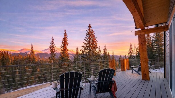 Una terraza con dos sillas Adirondack, una mesa y vistas a un bosque y montañas al atardecer.
