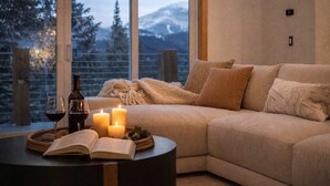 Una habitación acogedora con un sofá beige, una mesa redonda con un libro y una botella, velas, y vistas a montañas nevadas a través de amplios ventanales.