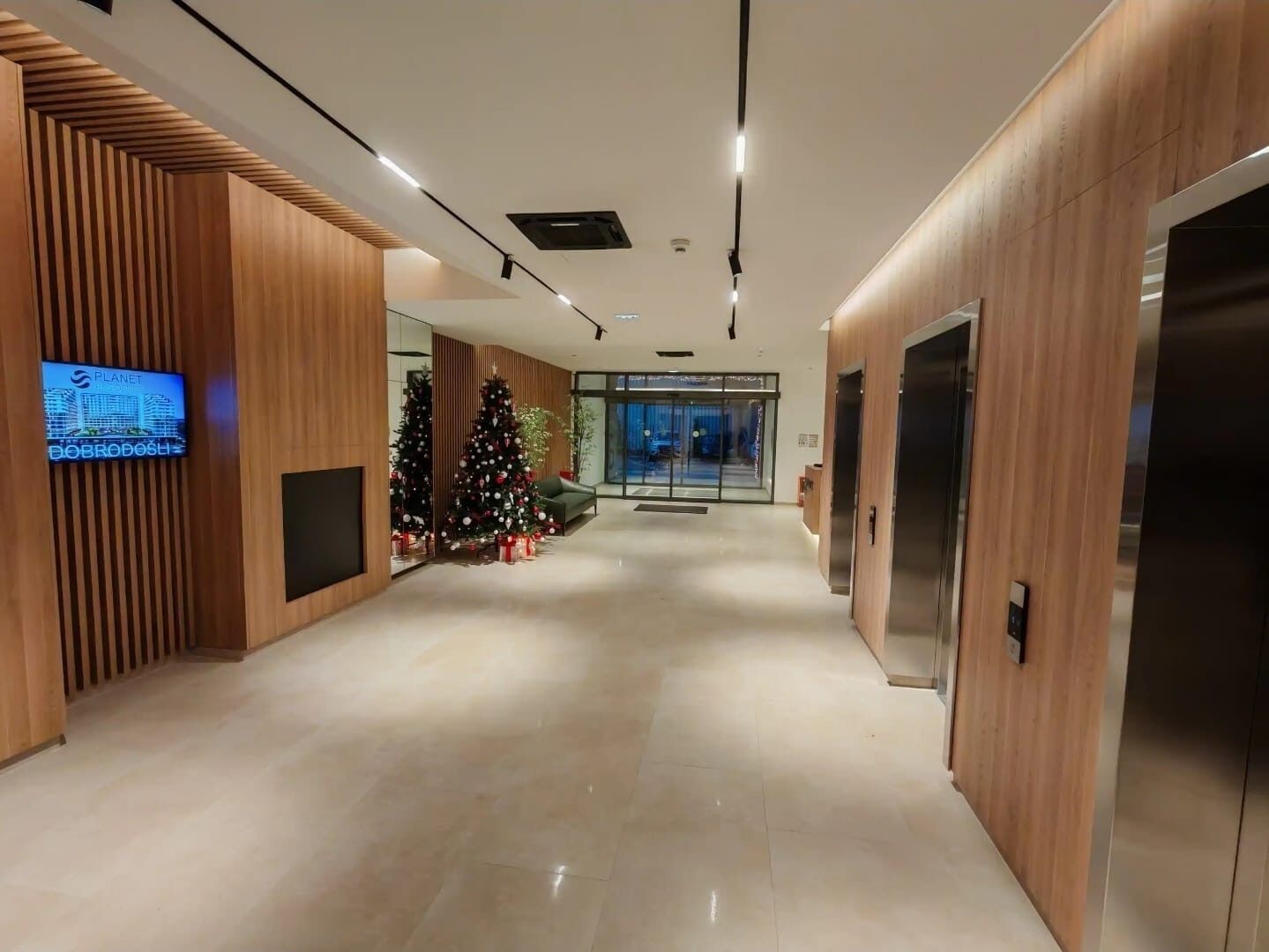 Un vestíbulo moderno con paneles de madera, una pantalla digital, dos árboles de Navidad decorados y una entrada de vidrio.