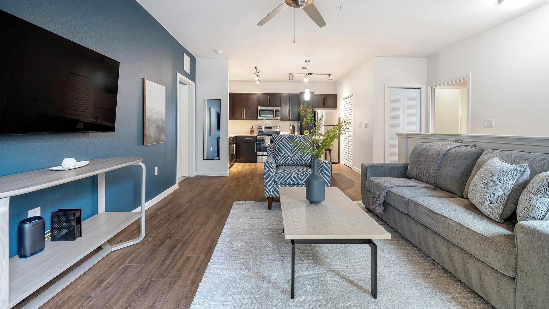 A modern living room with a flat-screen TV, a sofa, a coffee table, and a dining area with a kitchen in the background.