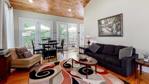 Un salon avec un plafond en bois, un canapé noir, un fauteuil beige, une table basse et un coin repas avec une table et des chaises.