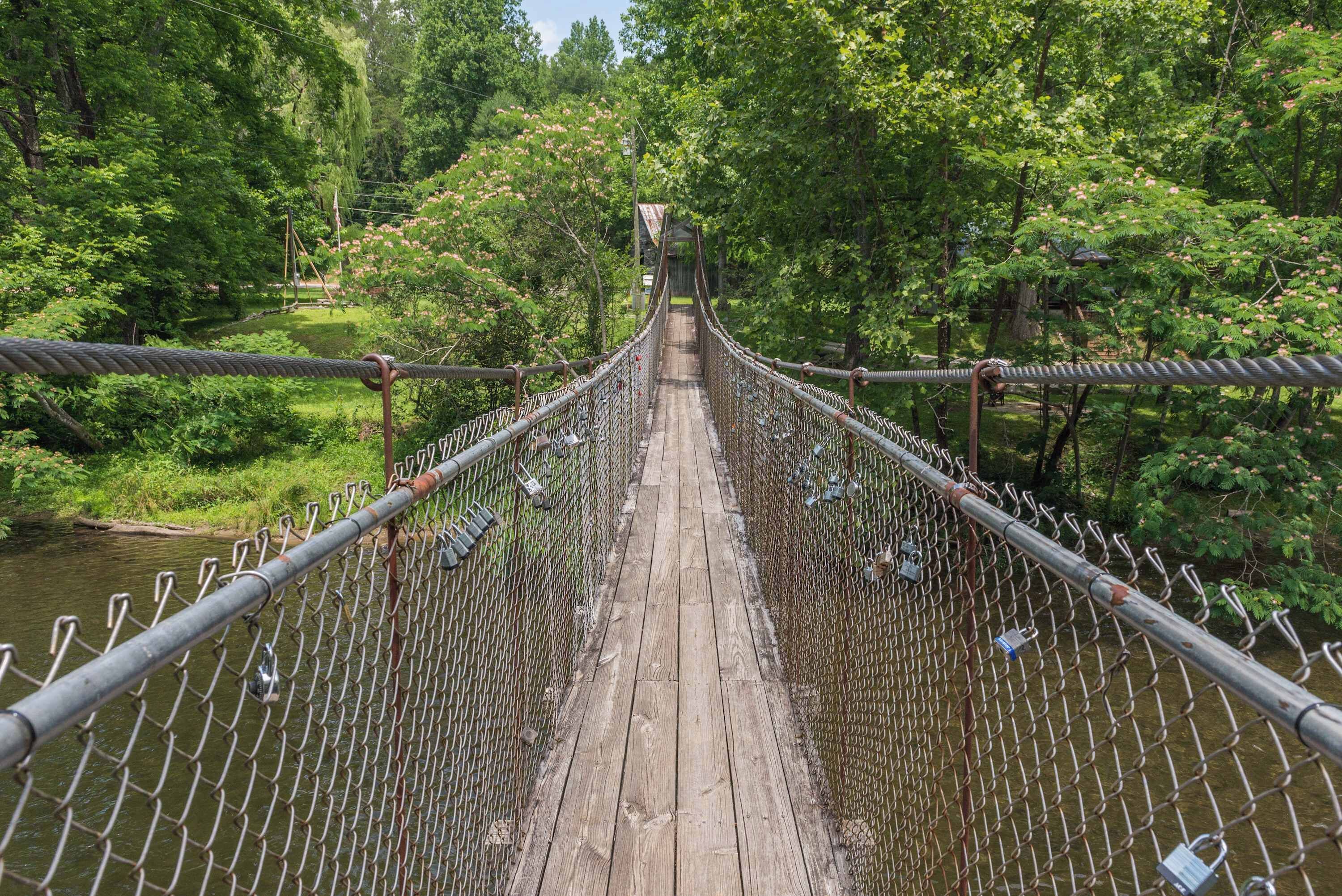 A suspension bridge with metal railings and wooden planks, surrounded by lush greenery and trees.