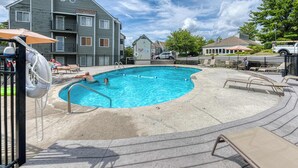 Un espace piscine avec des chaises longues et un poste de surveillance pour le sauveteur, entouré de bâtiments et d’un parking.