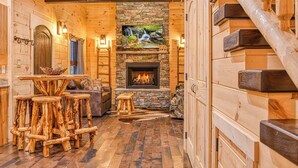 A log cabin interior with a stone fireplace, wooden furniture, and a staircase. - Creekside Luxury Cabin, Hot Tub, Near Downtown (Gatlinburg)