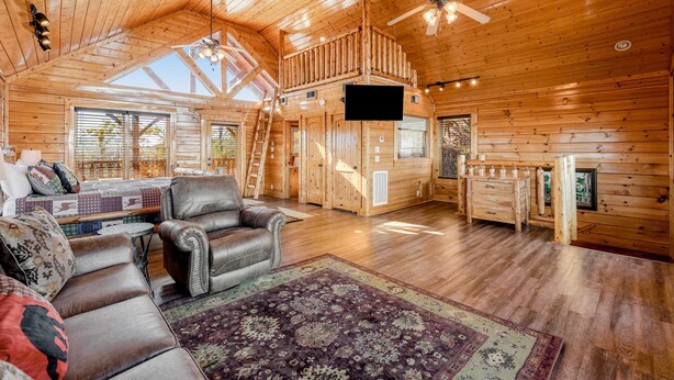 A spacious log cabin interior with a loft bedroom, a fireplace, a large window, and a leather sofa. - Mtn Retreat, for 12・Hot Tub・Games・Deck・Shared Pool (Sevierville)