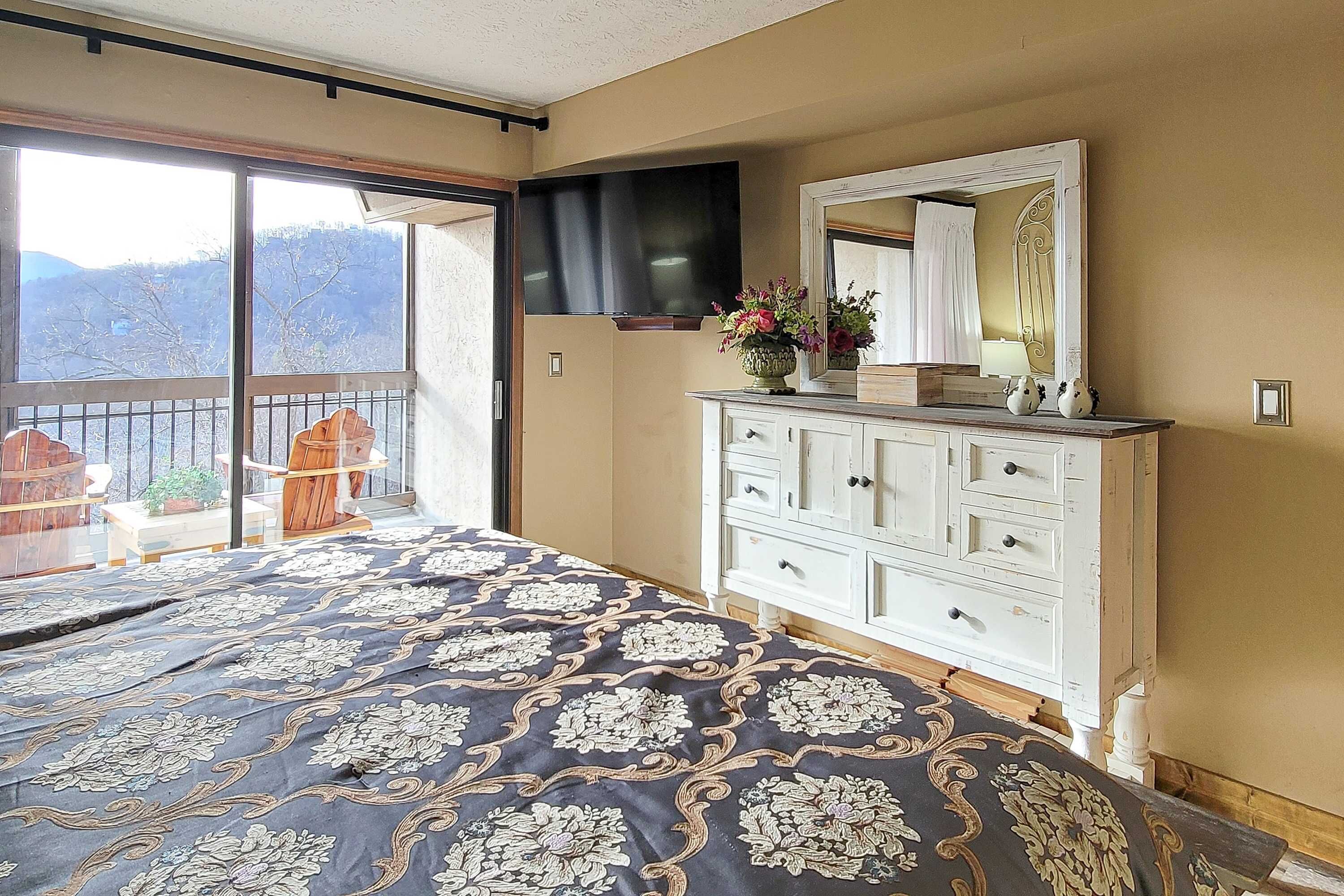 Un dormitorio con una cama grande, un aparador, un espejo y un televisor. Hay un balcón con vistas a árboles y colinas.