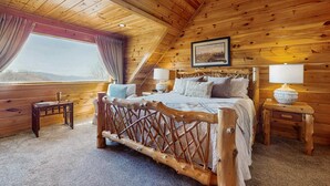 Ein Schlafzimmer einer Holzhütte mit einem Bett, zwei Nachttischen, einem Fenster mit Vorhängen, einem gerahmten Bild an der Wand und einem Deckenventilator.
