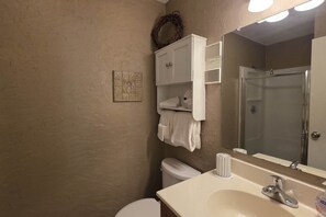 A bathroom with a beige textured wall, a white toilet, a sink with a mirror, and a shower enclosure. - Ski Mountain Condo | Stunning Views & Balcony (Gatlinburg)