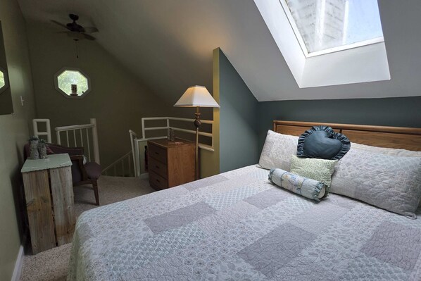 A bedroom with a skylight, a bed with patterned bedding, a bedside table with a lamp, and a wooden dresser. - Ski Mountain Condo | Stunning Views & Balcony (Gatlinburg)