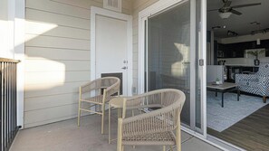 A covered patio area with two wicker chairs and a table, a sliding glass door leading to a kitchen area, and a ceiling fan. - Landing | Modern 2BD, Gym, Clubhouse (Cedar Park)