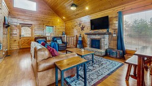 A log cabin interior with a wooden ceiling, a fireplace, a leather sofa, a television, and a large window with blinds. - Lovely Cabin, Sleep 6・Hot Tub・Mtn View・Pool Access (Sevierville)