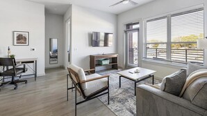 A modern living room with a sofa, armchair, coffee table, and a desk with a chair. There is a TV mounted on the wall, a window with blinds, and a door leading to another room. - Landing | Incredible 2BD, Gym, Pool (Plano)