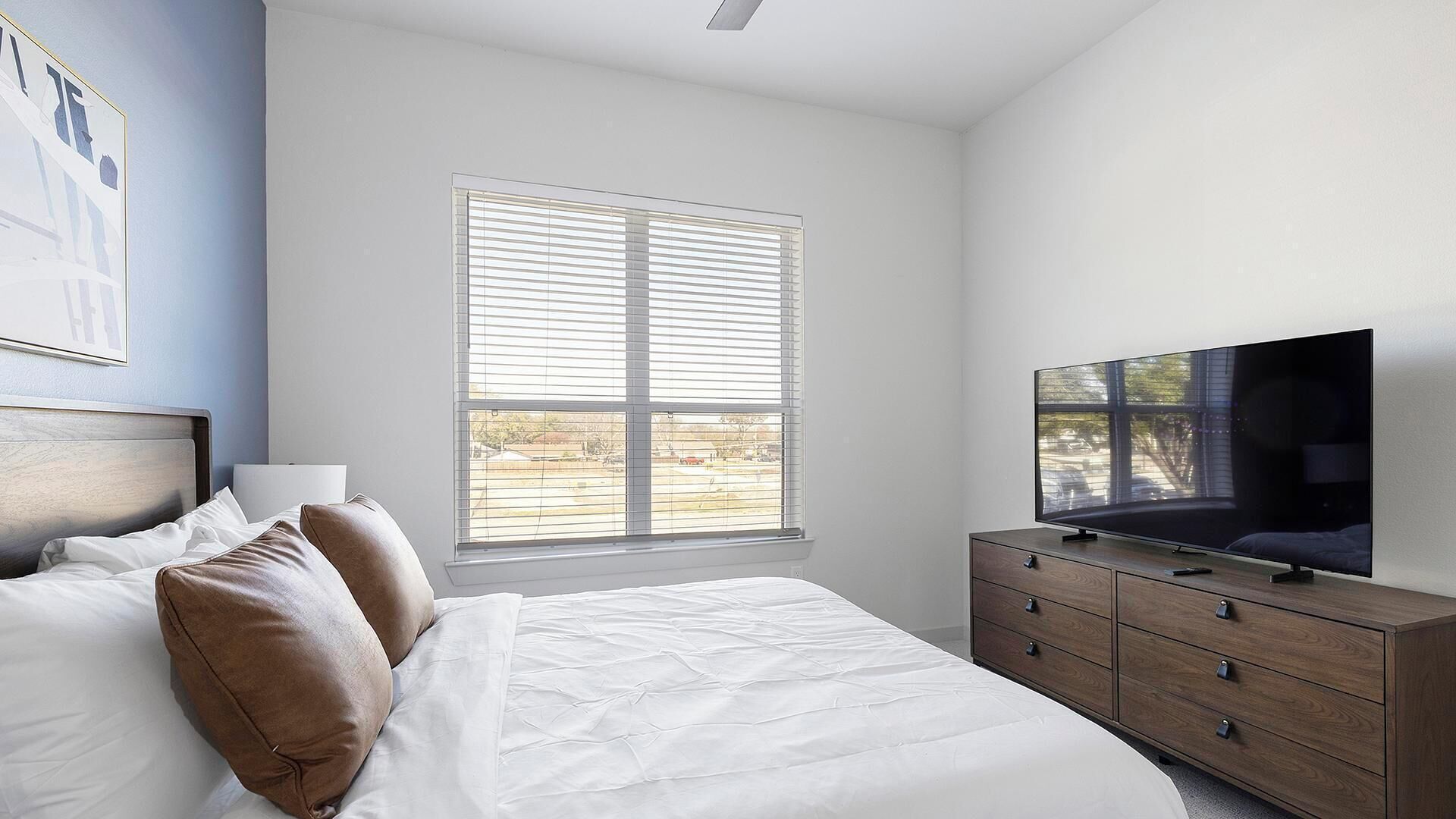Un dormitorio con una cama, televisor, cómoda y ventana con persianas.
