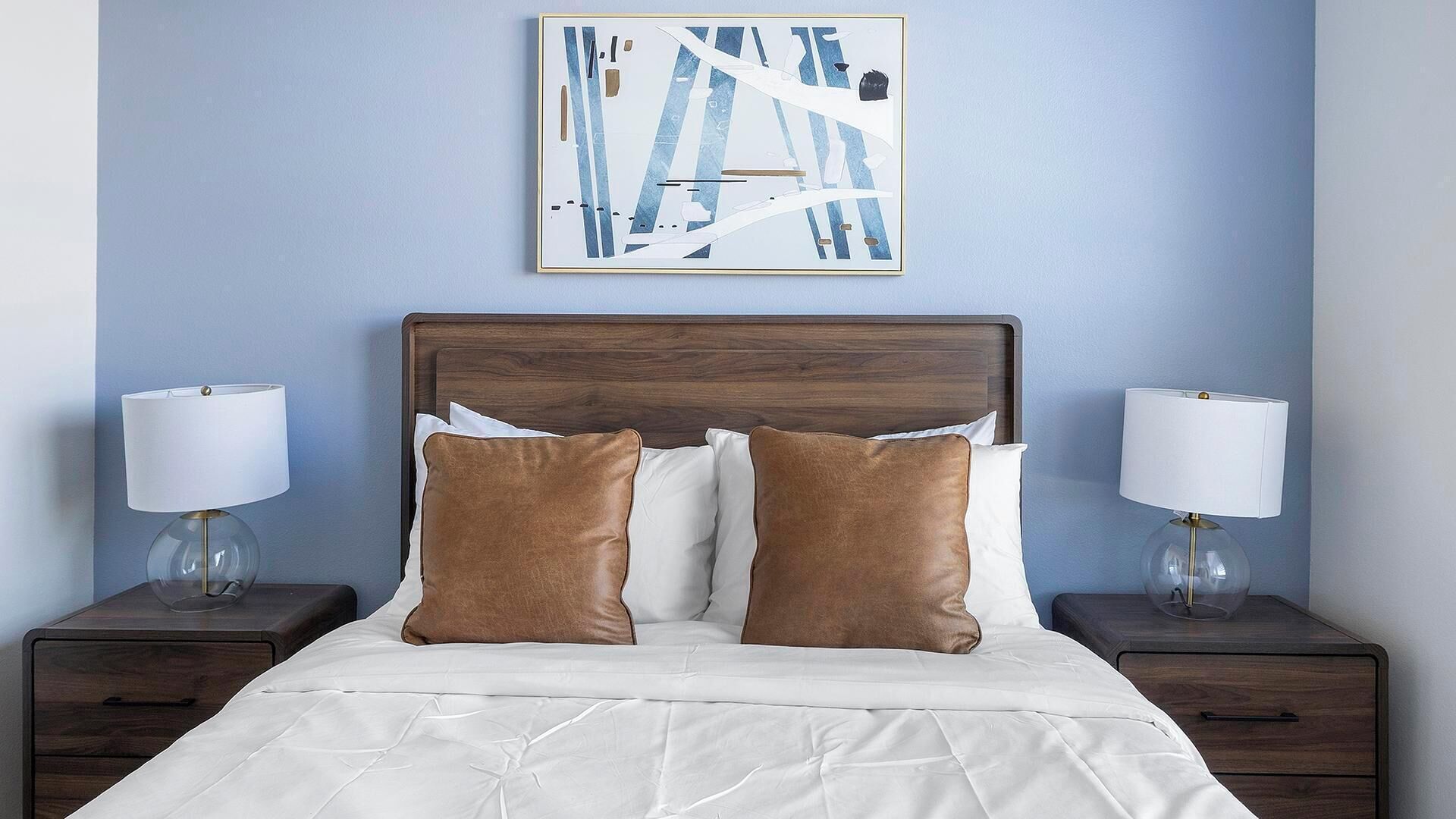 A neatly made bed with white bedding and two brown pillows, flanked by two bedside tables with lamps, against a blue wall with a piece of abstract art above.