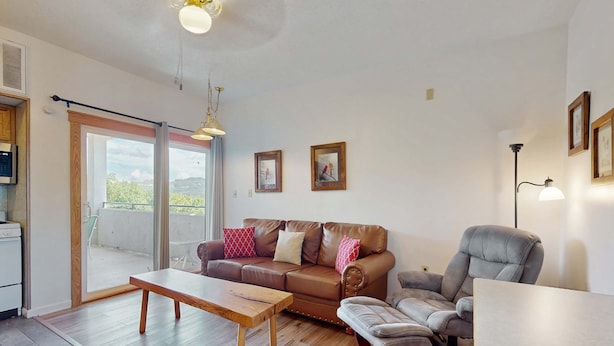 Ein Wohnzimmer mit braunem Sofa, einem grauen Sessel, einem Holztisch und einem Esstisch. Eine Glastür führt auf einen Balkon.