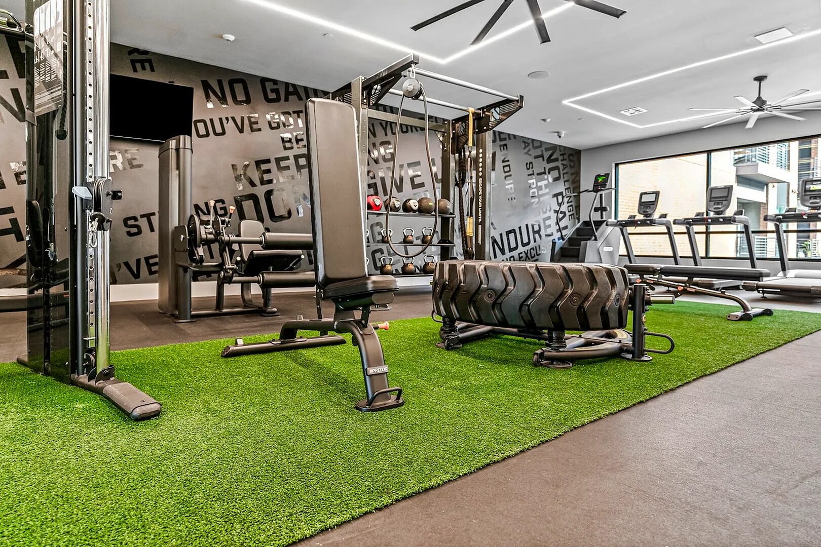 A gym with various exercise equipment, a green artificial turf floor, and motivational wall art.