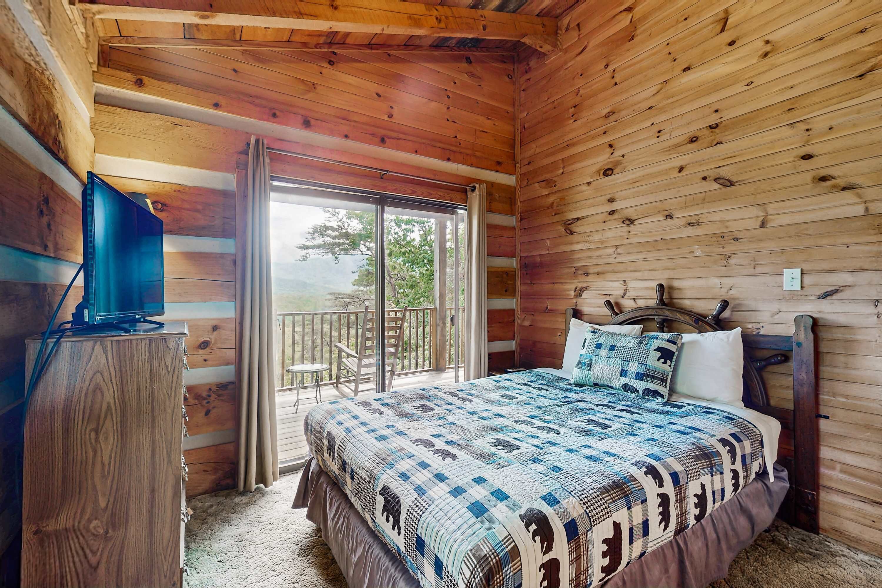 A wooden cabin bedroom with a bed, a television on a wooden cabinet, and a balcony with a chair.