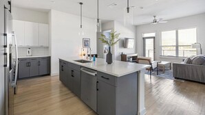 A modern kitchen with grey cabinets, a large island, and pendant lighting. - Landing | Spectacular 2BD, Pool, Gym (Plano)