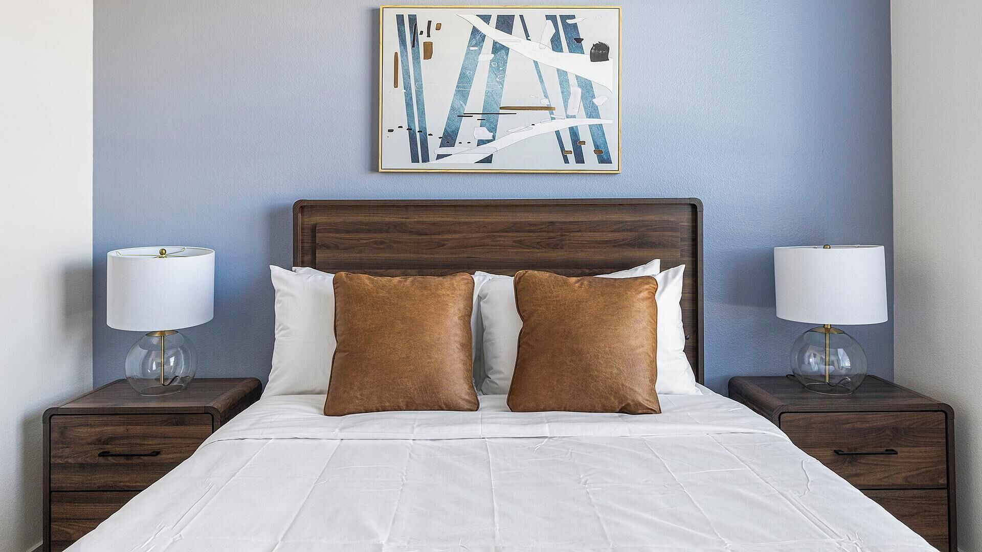 A neatly made bed with white linens and two brown pillows, flanked by two bedside tables with lamps, against a blue wall with a framed abstract painting above.